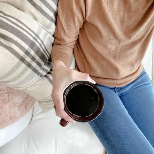 person in brown sweater sitting on white couch with coffee mug in hand